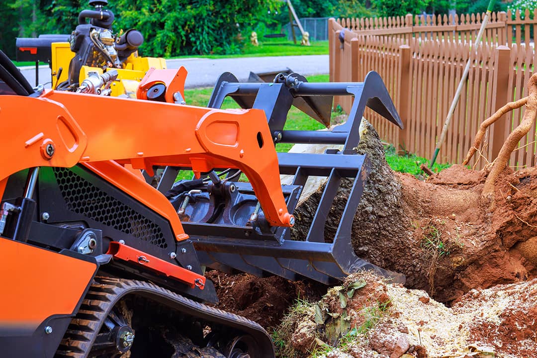 A compact track loader extracting large tree stump from a backyard in a suburban neighborhood.